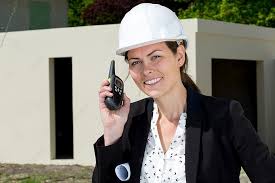 Female construction manager in a hard hat holding a radio at a building site, smiling confidently. Represents leadership in construction industry.