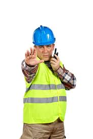 Construction worker in a blue hard hat and reflective vest, signaling with a hand raised while holding a walkie-talkie, emphasizing safety communication.