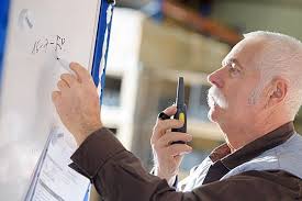 An older man with a white mustache uses a marker on a whiteboard while holding a two-way radio, indicating a work-related discussion or instruction.