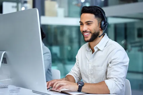 Smiling customer service representative in a headset, engaging with a computer at a modern office workspace. Demonstrates professionalism and support.