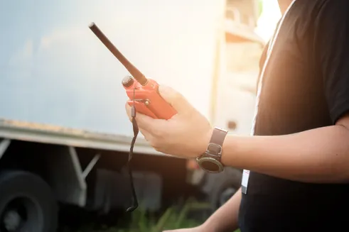 A person in a black shirt holds an orange two-way radio, standing near a truck. The image highlights communication in a logistical setting.